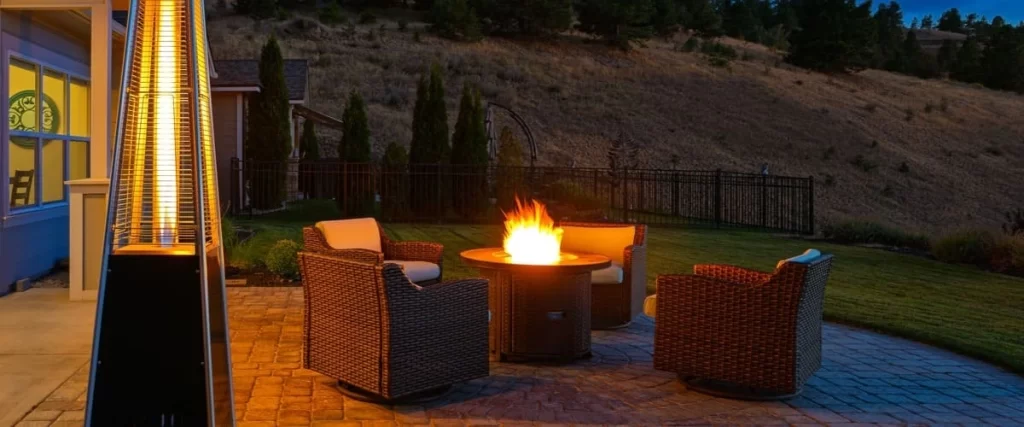 A cozy evening backyard patio setup featuring a circular gas fire pit table surrounded by dark wicker swivel chairs on a stone paver floor with a tall pyramid heater in the foreground.