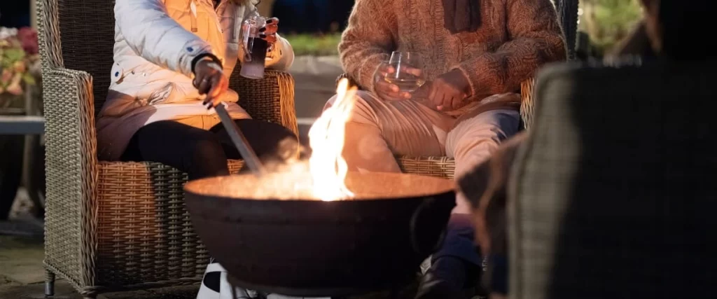 Two people sitting in wicker chairs enjoying drinks at night next to a glowing metal bowl fire pit with bright flames.