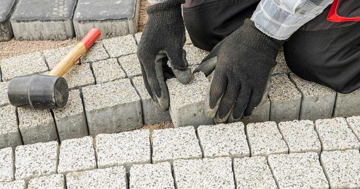 A close-up of a professional landscaper wearing black gloves carefully placing grey granite stone blocks into a curved pattern, demonstrating the use of the best patio materials for a durable outdoor living space.