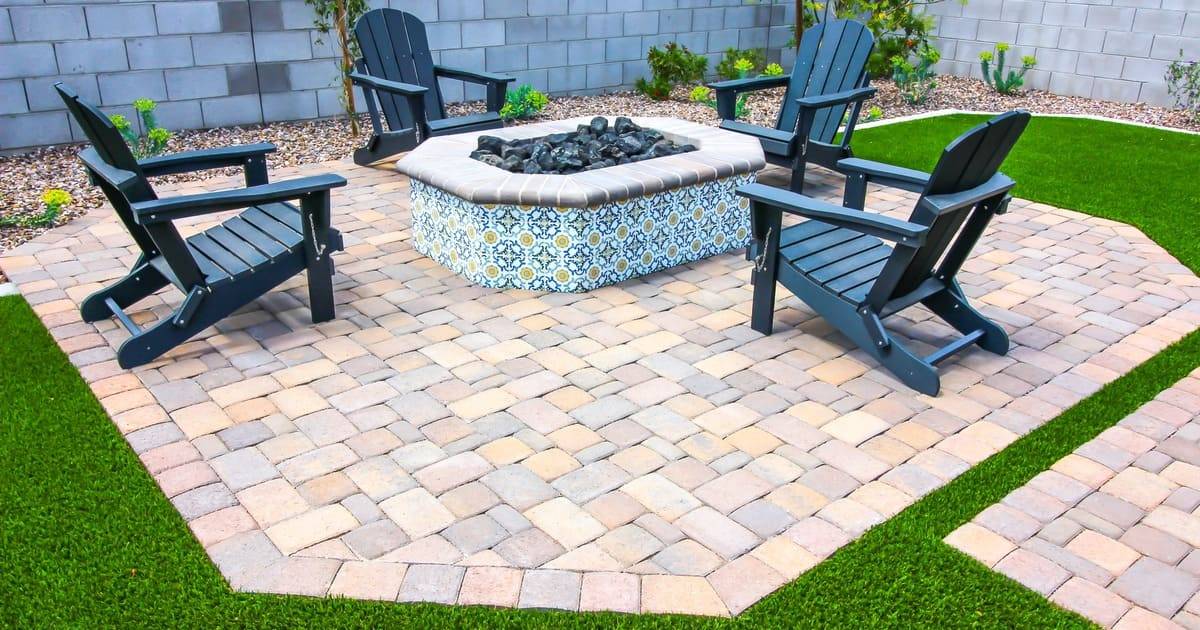 A beautifully maintained backyard patio featuring expert paver sealing on a stone walkway surrounding a tiled fire pit with blue Adirondack chairs.