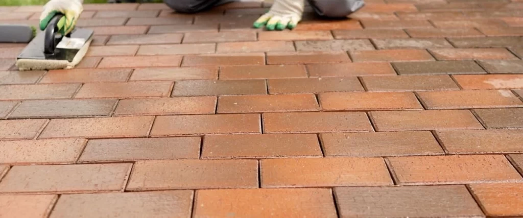 A contractor wearing protective gloves uses a specialized applicator tool to apply a clear protective sealant over red and brown brick patio pavers to prevent staining.