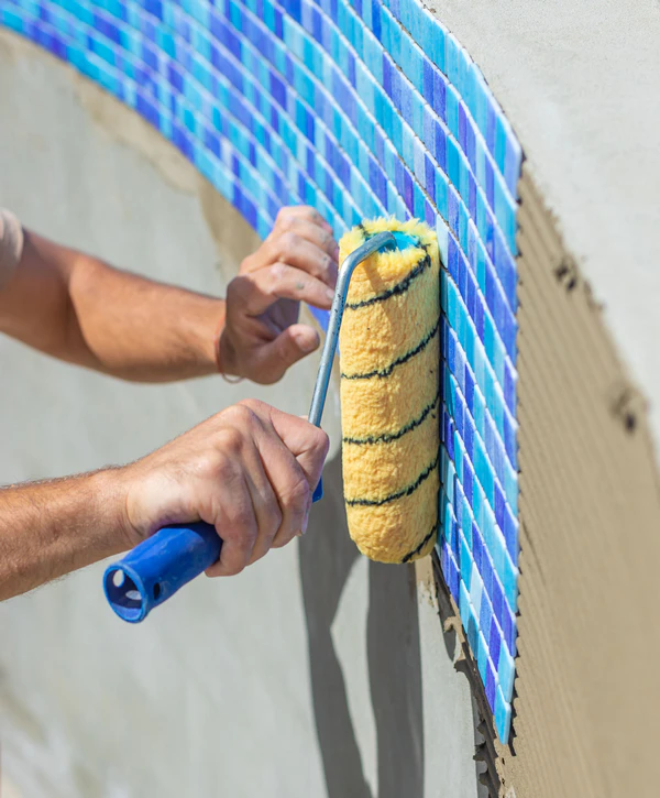 A contractor using a roller to apply adhesive for new blue mosaic tiles during a professional pool repair in Carrollton, TX.