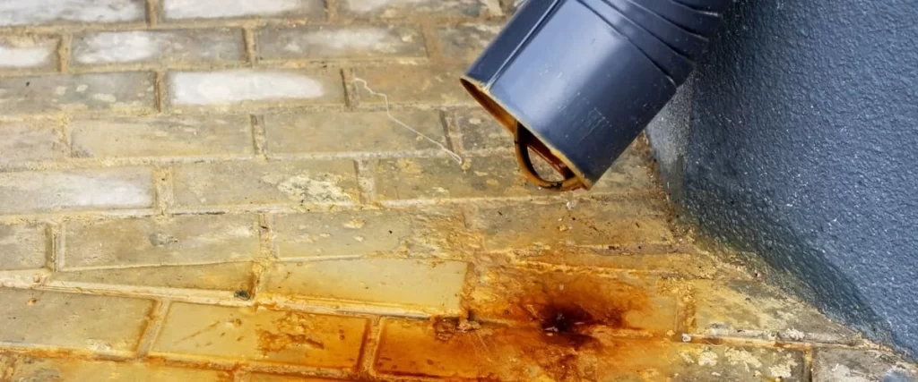 Close up view of orange rust stains and water mineral deposits accumulating on light-colored patio pavers located directly beneath a black downspout drainage pipe.