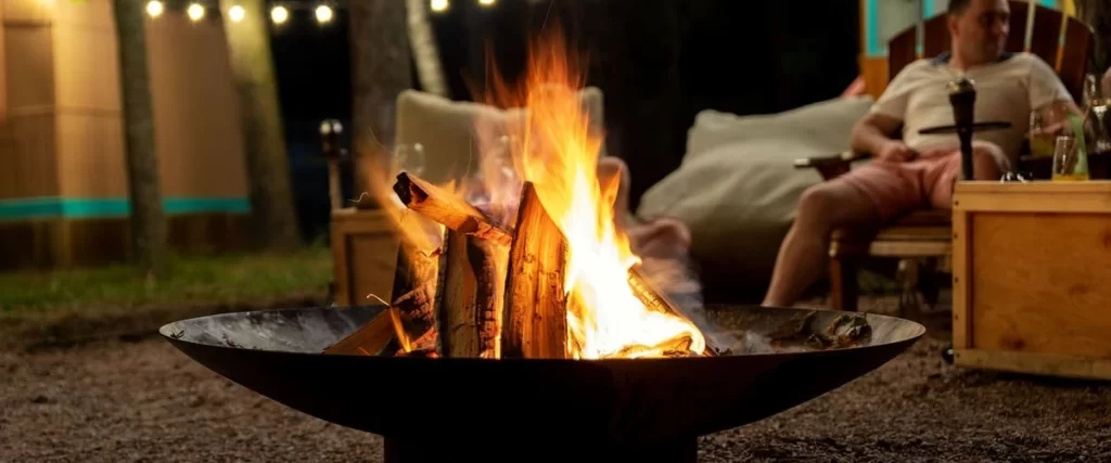 A large steel bowl fire pit burning logs in a backyard setting with decorative string lights and a person relaxing in the background.