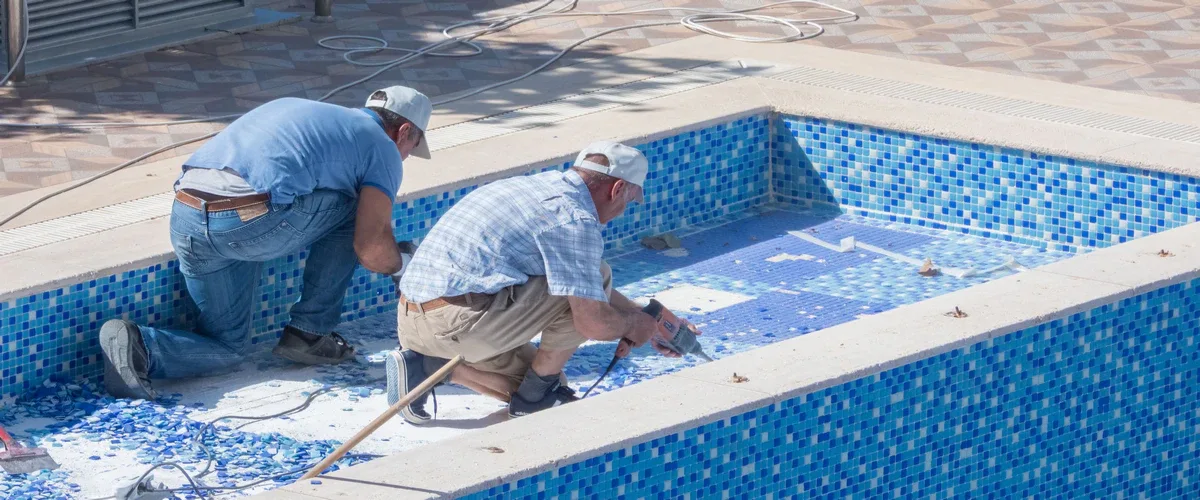 Two contractors working inside an empty blue mosaic tile swimming pool to perform a structural repair and surface restoration using power tools.