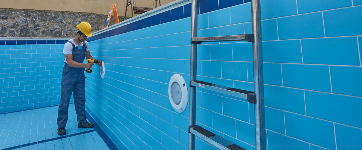A pool service professional in a hard hat and overalls using a power drill to perform a repair on an underwater LED light fixture inside an empty blue tiled swimming pool.