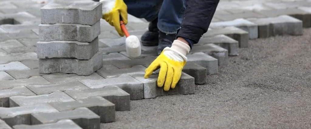 A worker in yellow safety gloves laying grey I-shaped interlocking concrete pavers on a leveled gravel surface.