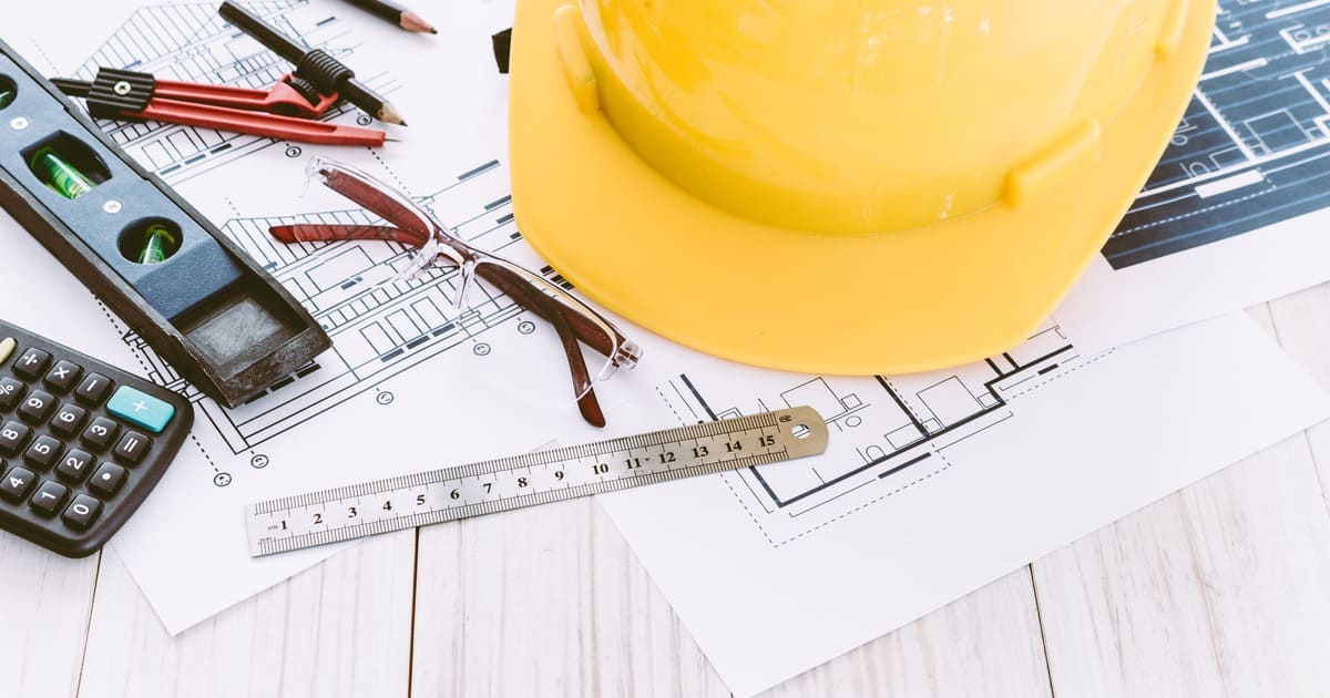 An overhead shot of a bright yellow construction hard hat resting on top of architectural plans, surrounded by a calculator, eyeglasses, a spirit level, and a compass.
