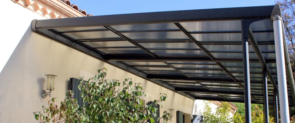 A dark grey aluminium pergola attached to a house wall featuring translucent roof panels for weather protection.