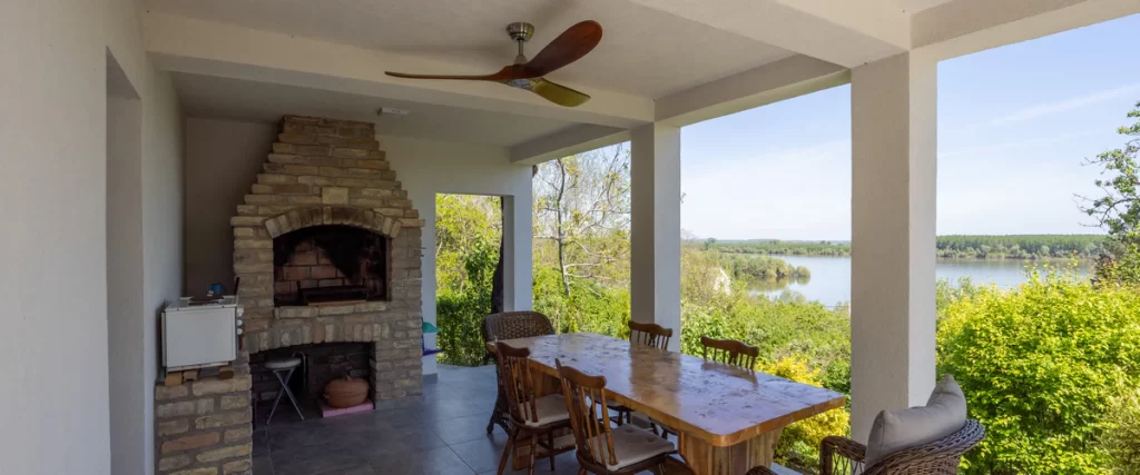 A large wooden-blade ceiling fan installed in a covered outdoor patio area featuring a brick fireplace and dining table.