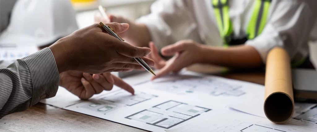 Professionals pointing at architectural blueprints on a desk to ensure the new pergola meets building permit requirements.