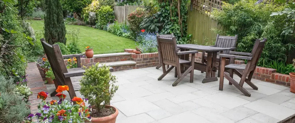 A peaceful and cool patio featuring stone pavers, wooden garden furniture, and lush green landscaping for summer shade.