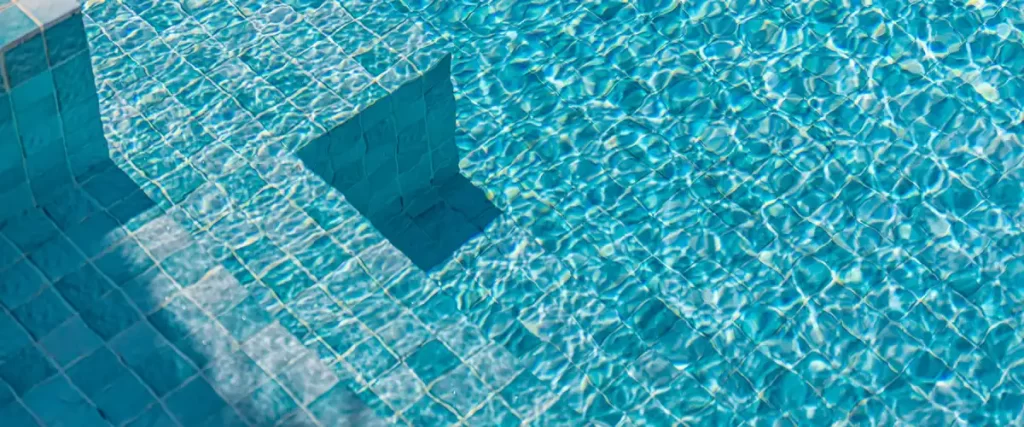 Sunlight reflecting off the ripples of crystal clear water in a swimming pool with bright blue mosaic floor tiles.