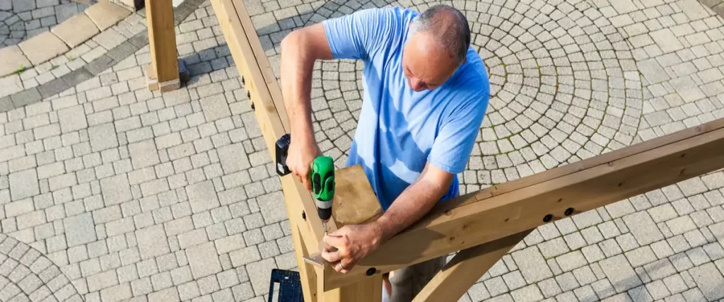 A man using a green power drill to secure wooden beams during a weekend DIY backyard pergola project on a paved patio.