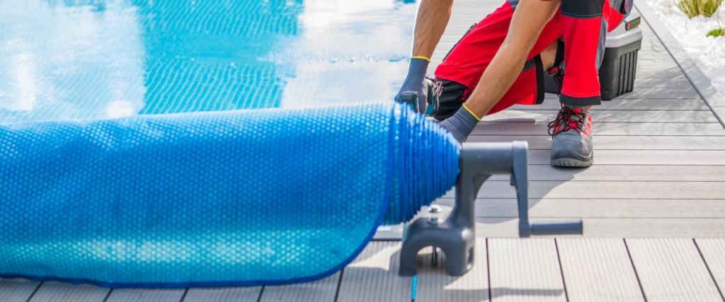 Close up of a worker installing a blue bubble-style pool cover near textured aggregate finishes on a modern wooden deck.