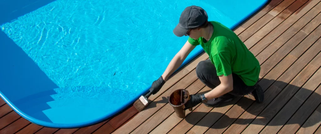 A person in a green shirt and cap carefully applying brown stain to a wooden deck bordering a blue swimming pool.