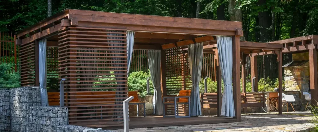Luxury wooden garden pergola featuring light grey outdoor privacy curtains and slatted walls.