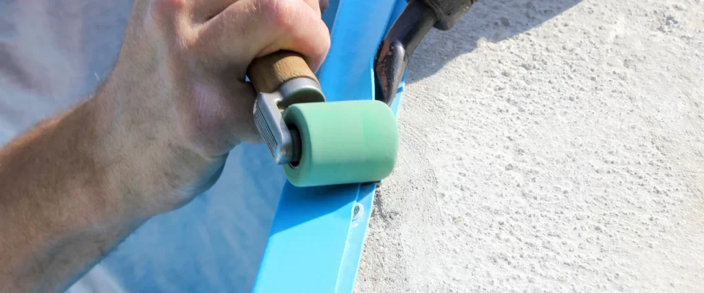 A close-up view of a technician's hand using a green silicone pressure roller to smooth and secure a bright blue pool liner against a concrete surface.