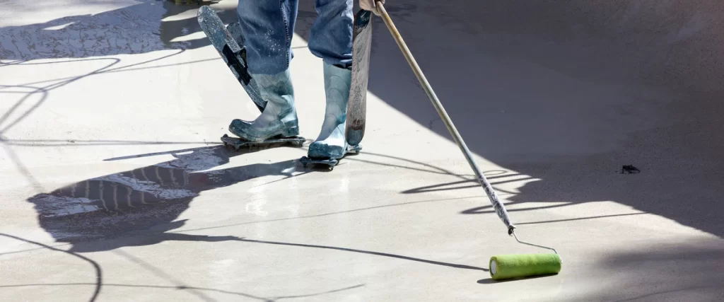 A contractor wearing spiked boots uses a roller to apply a smooth white pool and plaster finish to the interior surface.