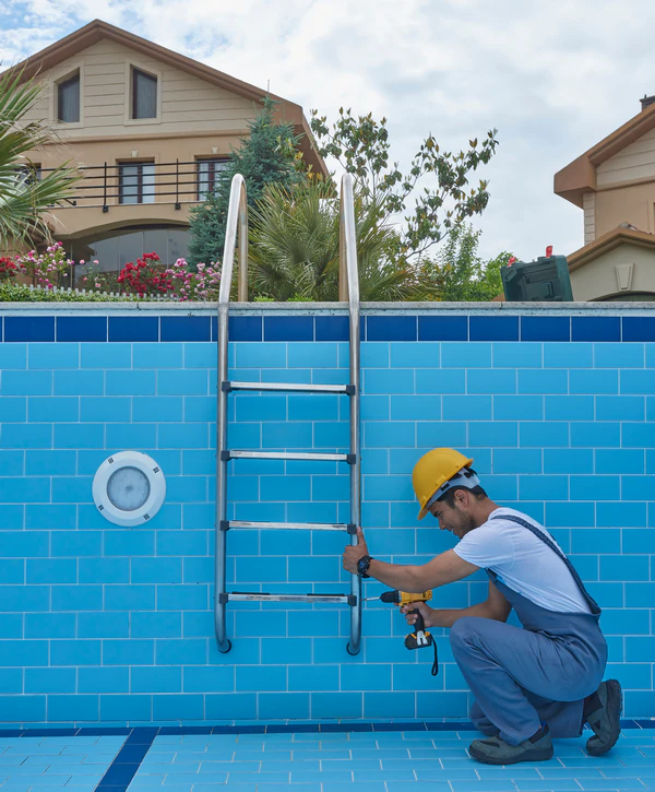 A pool repair expert using a power drill to secure a stainless steel ladder to the blue tiled wall of a backyard swimming pool in Lewisville, TX.