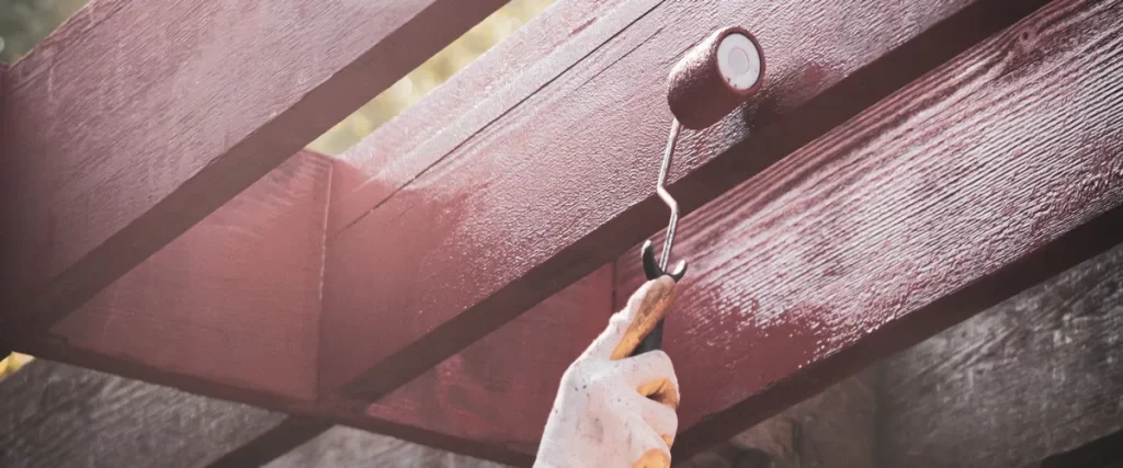 A person wearing a glove using a paint roller to apply a dark reddish-brown stain during routine pergola maintenance.