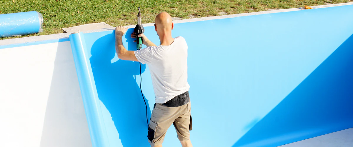 A technician performing a liner repair by using a specialized heat welding tool to seal a new blue vinyl membrane to the side of a swimming pool.
