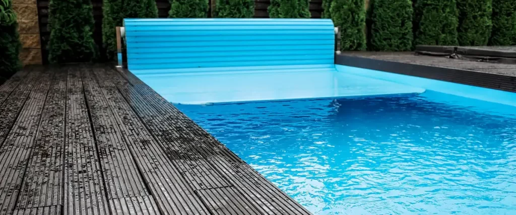 A bright blue slatted automatic pool cover partially extended over the water of a backyard swimming pool adjacent to a wet wooden deck.
