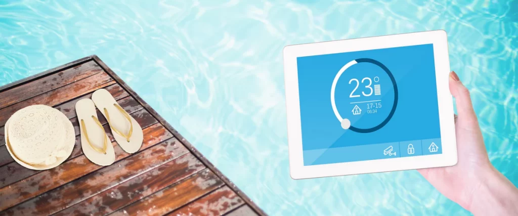A hand holding a digital tablet displaying a smart pool automation system interface showing a 23-degree temperature, with a sunny pool and wooden deck in the background.
