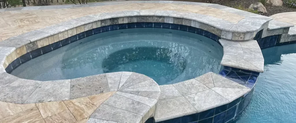 A custom built-in pool spa featuring elegant grey stone coping, blue waterline tiles, and submerged seating integrated into a larger swimming pool.