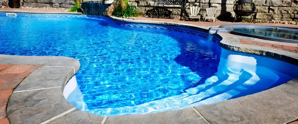 A tropical-style home swimming pool featuring decorative water fountains, stone tile decking, and surrounding garden greenery.