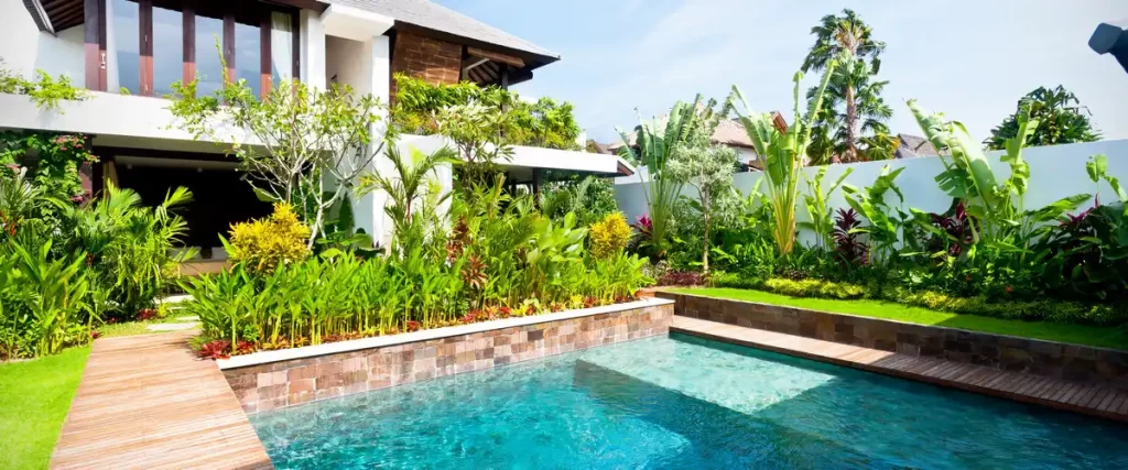 A stunning tropical pool landscape featuring vibrant green broadleaf plants, manicured grass, a wooden walkway beside a clear pool, and a modern home exterior.