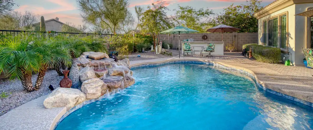An elegant freeform swimming pool with a custom stacked stone waterfall feature, desert-style landscaping, and an outdoor kitchen bar in the background.