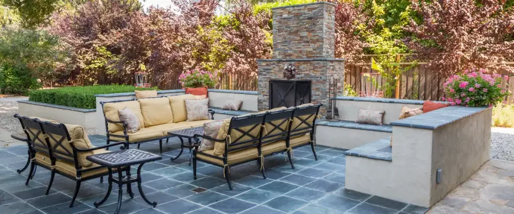 A luxury outdoor living space featuring a custom stone fireplace, slate tile flooring, and built-in concrete seating with yellow cushions.