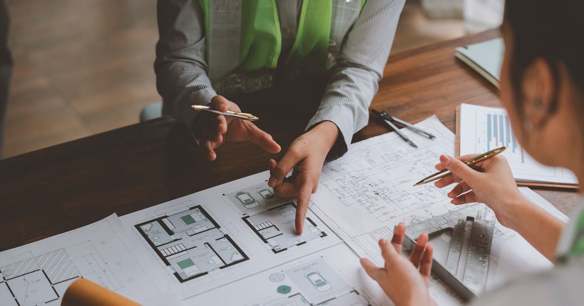 Close-up of an architect and client discussing detailed floor plans and blueprints on a wooden desk during a home renovation meeting.