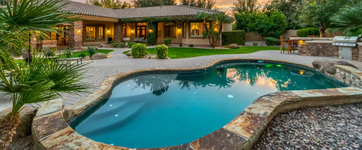 A wide view of a large Mediterranean-style home at dusk featuring a curvy swimming pool with stone edging, a spacious paved patio, and manicured green lawns.