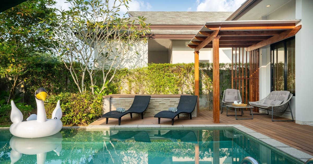 A beautifully renovated backyard featuring modern pool upgrades including a wooden deck, shaded pergola with lounge chairs, tropical landscaping, and a swan floatie.