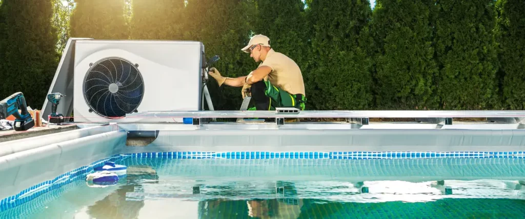 A professional technician in uniform connecting wiring to an electric pool heater and heat pump installed next to a backyard swimming pool.
