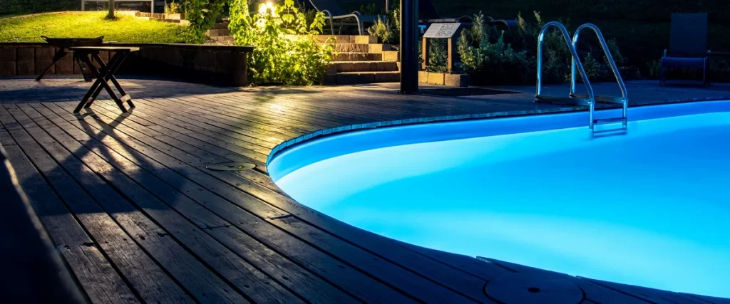 A backyard swimming pool beautifully illuminated at night by bright blue LED pool lighting, surrounded by a dark wooden deck and landscape lights.