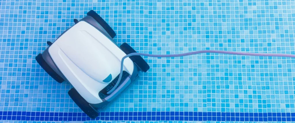Top-down view of a white automatic robotic pool cleaner moving across a light blue mosaic tiled swimming pool floor connected to a floating cord.