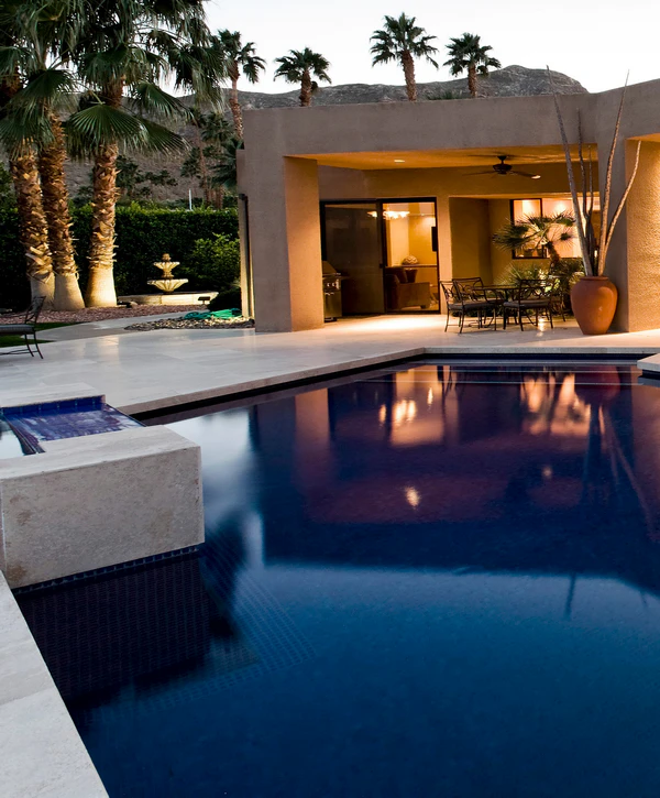 A luxurious dark blue swimming pool with an elevated stone spa at a modern home with palm trees at twilight, built by a premier pool designer in Little Elm, TX.