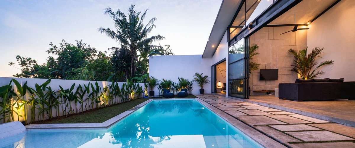 A sleek rectangular pool surrounded by tropical plants and a paved patio, seamlessly connecting to a modern open-concept white home at dusk.