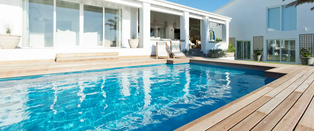 A rectangular swimming pool with clear blue water, surrounded by a custom wooden deck, situated immediately behind a modern white home featuring large glass sliding doors and a covered patio.