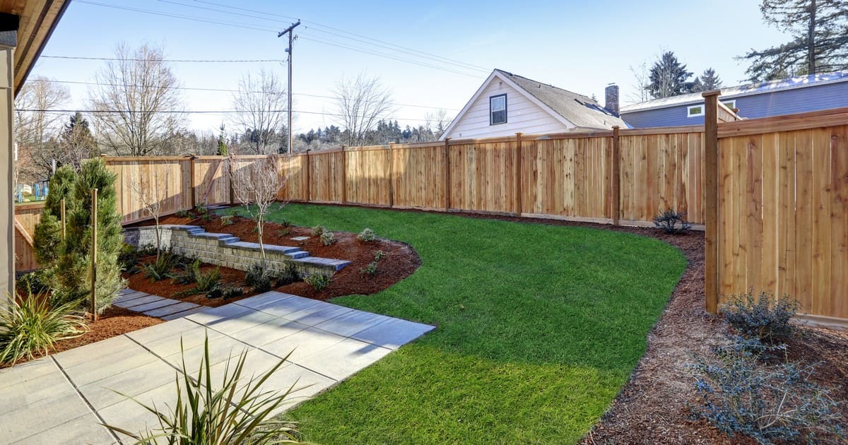 A newly landscaped backyard featuring a tall wooden privacy fence, fresh green grass, a small stone retaining wall with garden plants, and a concrete patio area.