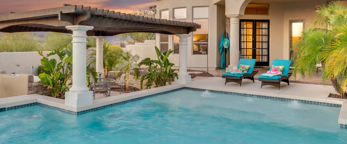 Beautiful light blue swimming pool with small water fountain jets, situated next to a patio with a white column pergola and bright blue lounge chairs at sunset.