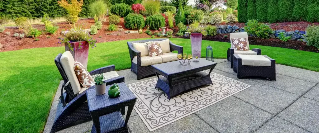 A comfortable outdoor living space featuring a dark wicker patio furniture set on a concrete pad, surrounded by a beautifully landscaped backyard with green grass and garden beds.