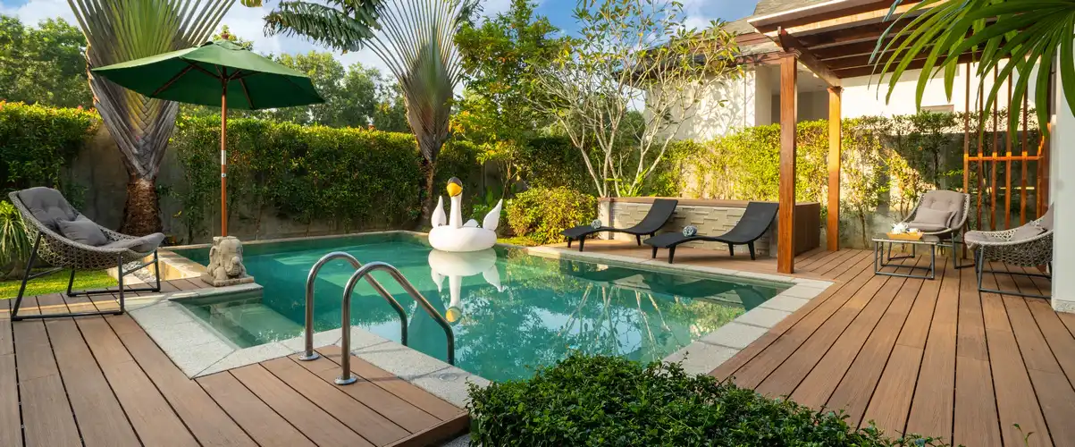 Small rectangular swimming pool surrounded by a wooden deck, featuring a white swan float, lounge chairs, and a green umbrella in a lush tropical backyard.