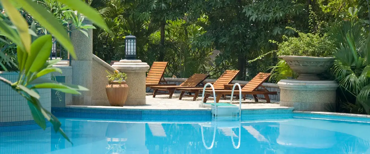 A clear blue swimming pool surrounded by lush tropical greenery, decorative stone pillars with a lantern, and a row of wooden lounge chairs.