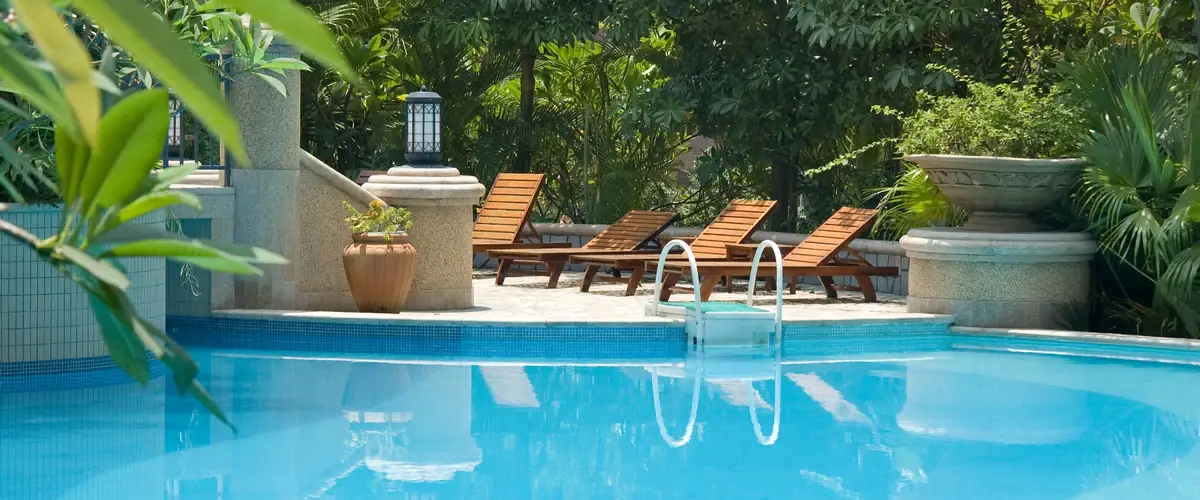 A bright blue swimming pool surrounded by a light stone patio, lined with four wooden sun loungers, and enclosed by lush, tropical green plants and landscaping.