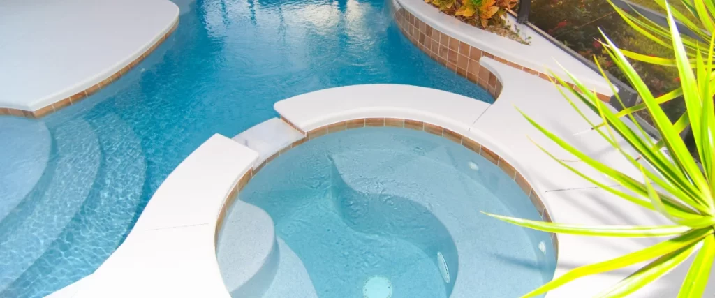 A close-up view of a curvy swimming pool design featuring a raised circular spa with a spillover edge and a separate curved tanning ledge area with shallow submerged steps.