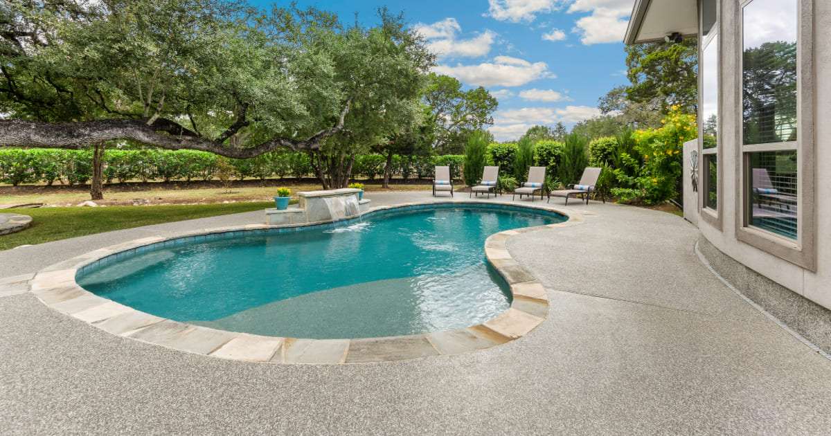 A freeform backyard swimming pool featuring a shallow entry area, showcasing practical pool tanning ledge ideas alongside a small stone waterfall feature and surrounding trees.